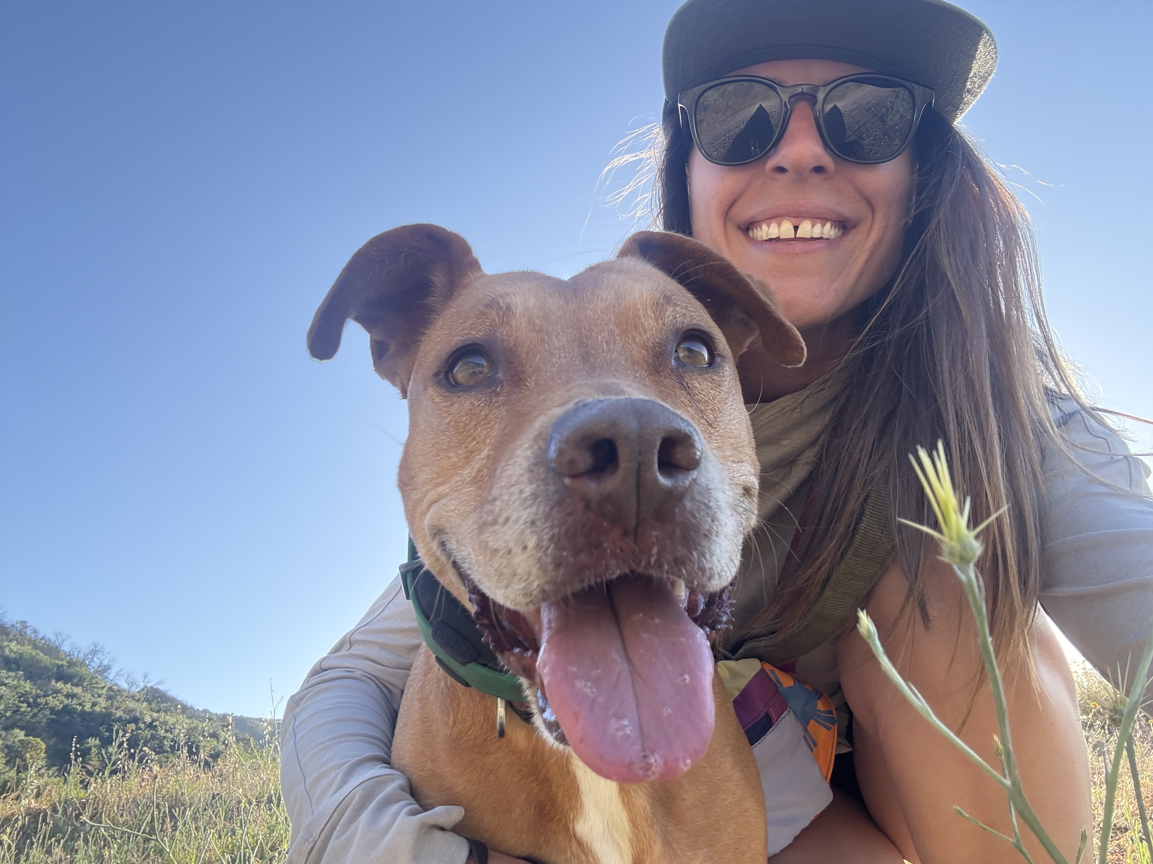 A smiling woman wearing a green hat crouches down next to a brown dog for a selfie. The dog is looking towards the camera with it's tongue out.
